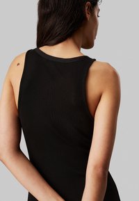 Top in cotone nero a costine con scollo rotondo e spalline larghe, che mostra il retro con una lentiggine visibile sulla spalla. Tessuto liscio, forma aderente.