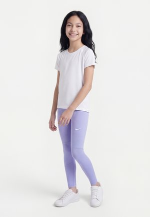Fille debout et souriante, portant une chemise blanche à manches courtes, des leggings Nike lavande et des baskets blanches sur un fond clair uni.