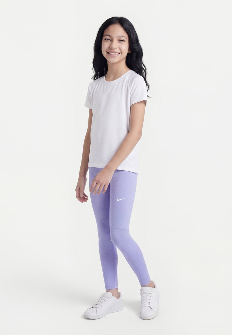 Fille debout et souriante, portant une chemise blanche à manches courtes, des leggings Nike lavande et des baskets blanches sur un fond clair uni.