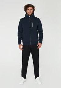 Sudadera azul marino con cremallera y cuello alto, bolsillo frontal y detalle de logo. Combinada con pantalones negros y zapatillas blancas. Textura de tela suave.