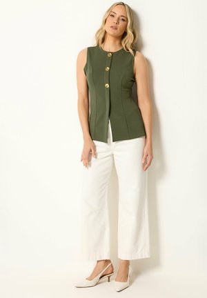 Mujer que lleva un top sin mangas verde oliva con botones, pantalones blancos de pierna ancha y tacones slingback blancos, de pie frente a un fondo claro.