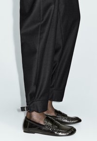 Black slip-on shoes with a glossy finish and subtle texture, paired with tailored black trousers featuring a cuff and metal adjustment detail.