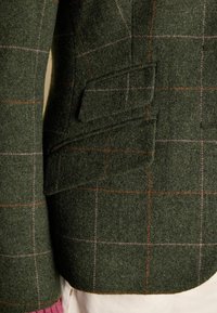 Tummanvihreä villatakki, jossa on ruudullinen kuvio oranssilla ja vaaleanpunaisella. Sisältää sivutaskun ja tekstuuripintaa. Lähikuva.