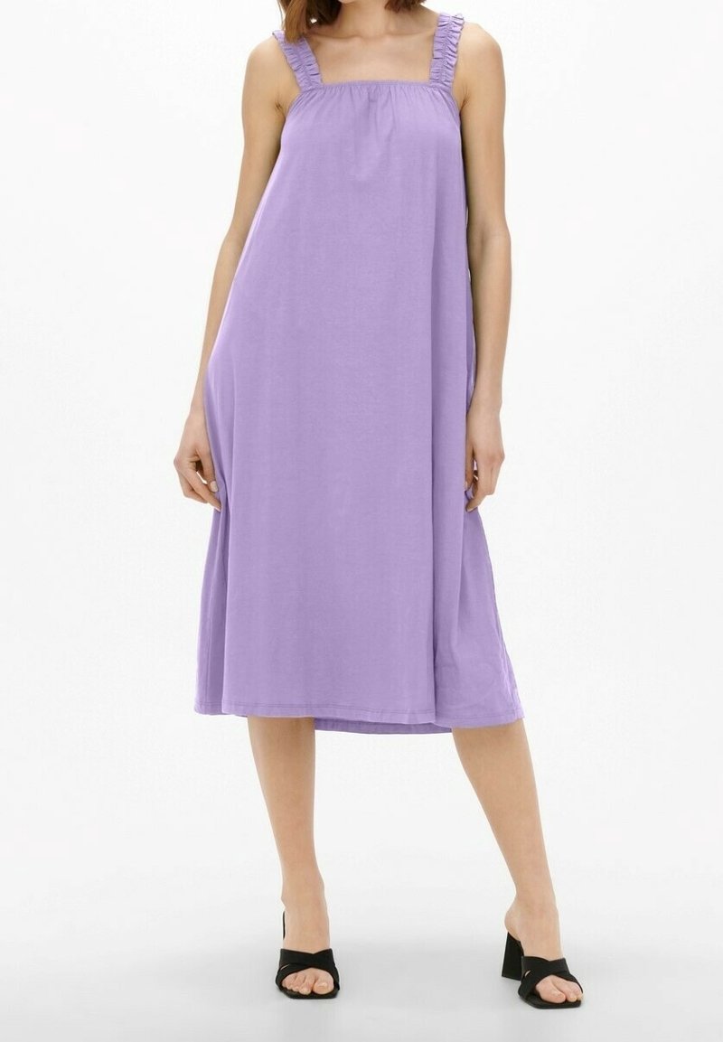 Femme portant une robe ample lavande longueur genou avec des bretelles à volants et des sandales à talons noires sur fond blanc.
