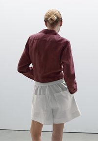 Bordeauxkleurig overhemd met kraag van zachte stof, gecombineerd met crèmewitte shorts met elastische taille en twee achterzakken.