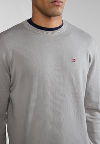 Suéter de punto gris claro con cuello redondo, detalles de canalé y un pequeño bordado de bandera roja y blanca en el lado izquierdo del pecho.