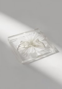 Klar fyrkantig akryldisplay som visar en vit blomma med detaljerade kronblad och två silverfärgade fjärilsaccent ovanpå.