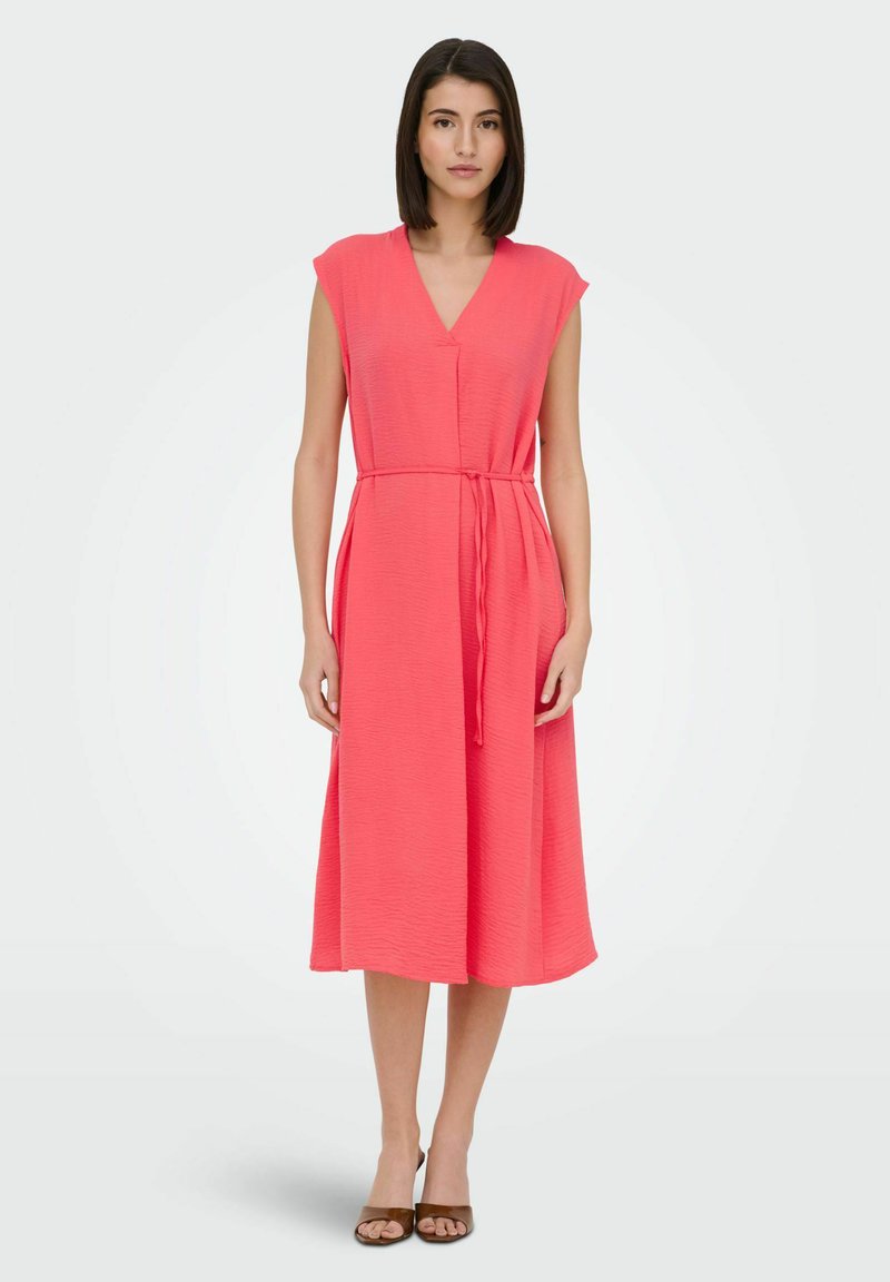 Femme debout portant une robe midi corail sans manches avec un col en V et une ceinture nouée à la taille, associée à des talons ouverts marron.