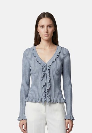 Donna che indossa un cardigan azzurro chiaro a coste con volant lungo la scollatura a V, bottone davanti, polsini e orlo, abbinato a pantaloni bianchi.