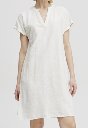 Donna che indossa un semplice abito bianco in lino con maniche corte, scollo a V e vestibilità rilassata su uno sfondo neutro.