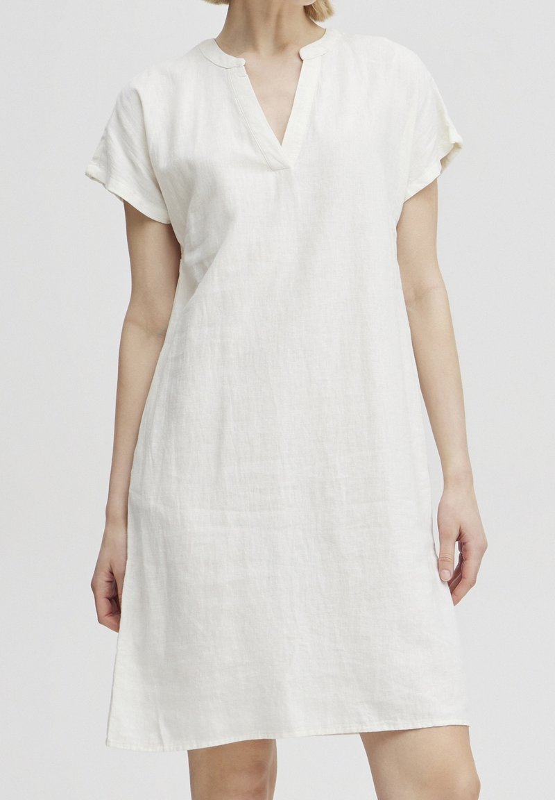 Femme portant une robe en lin blanche unie à manches courtes, col en V et coupe décontractée sur un fond neutre.