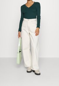 Person som bär en mörkgrön långärmad tröja och vida vita byxor, håller en ljusgrön handväska och har på sig beige plattformsskor.