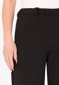 Pantaloni neri a vita elastica con una texture liscia. La vita è dotata di un cordoncino. Dettaglio della cucitura laterale visibile lungo la gamba.