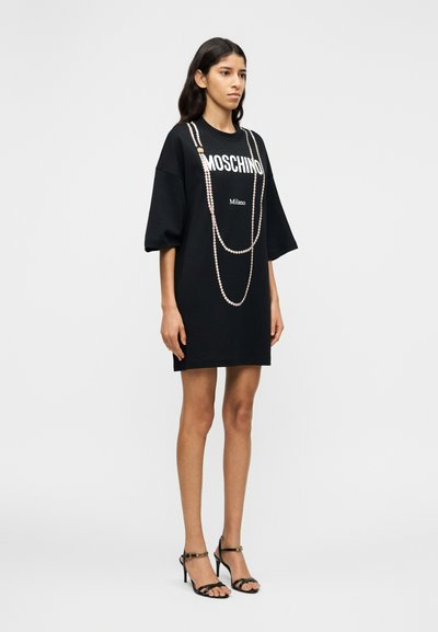 Μαύρο φόρεμα t-shirt με λευκό λογότυπο «MOSCHINO Milano» και διακοσμητική αλυσίδα που μοιάζει με πέρλες. Κοντά μανίκια και άνετη γραμμή. Μαύρα σανδάλια με λουράκια.