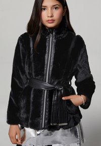 Jachetă din blană neagră cu guler înalt, fermoar frontal și curea din piele. Are o textură moale și o fustă lucioasă, metalizată, dedesubt.