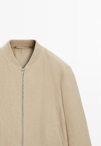 Beżowa kurtka bomberka z prążkowanym kołnierzem, srebrnym zamkiem błyskawicznym z przodu oraz otwarciami na kieszenie po bokach, przedstawiona na białym tle.