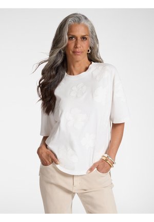 Donna con lunghi capelli grigi che indossa una maglietta bianca a maniche corte con motivi floreali, pantaloni beige e braccialetti oro e argento, con le mani nelle tasche.