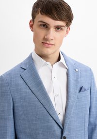 Helles blaues Blazer mit strukturiertem Gewebe, Reverskragen und einer Brusttasche. Getragen über einem weißen Hemd mit Knopfdetails.