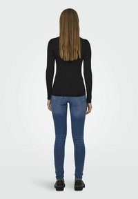 Svart langermet topp sammen med blå smale jeans og svarte sko med kraftig såle, sett fra bak mot en lys bakgrunn.