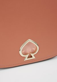 Gros plan d'une surface en cuir clair avec un emblème en forme de pique en or et cuir, avec des coutures visibles le long du bord.