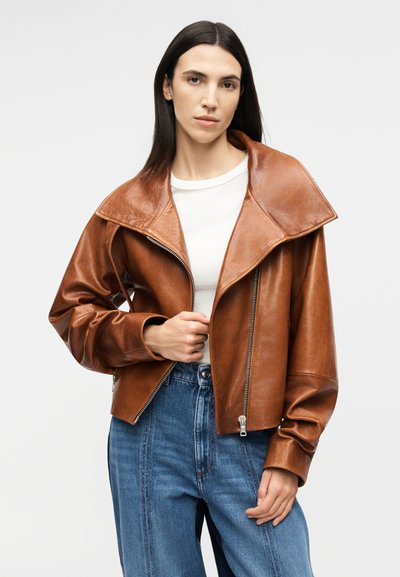 Γυναίκα που φοράει καφέ δερμάτινο μπουφάν πάνω από λευκό πουκάμισο και μπλε τζιν ψηλόμεσο, στέκεται μπροστά σε απλό φωτεινό φόντο.