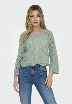 Lichtgroen gebreid vest met een golvende zoom en driekwart belmouwtjes, met een openwerk ontwerp tegen donkerblauwe jeans.