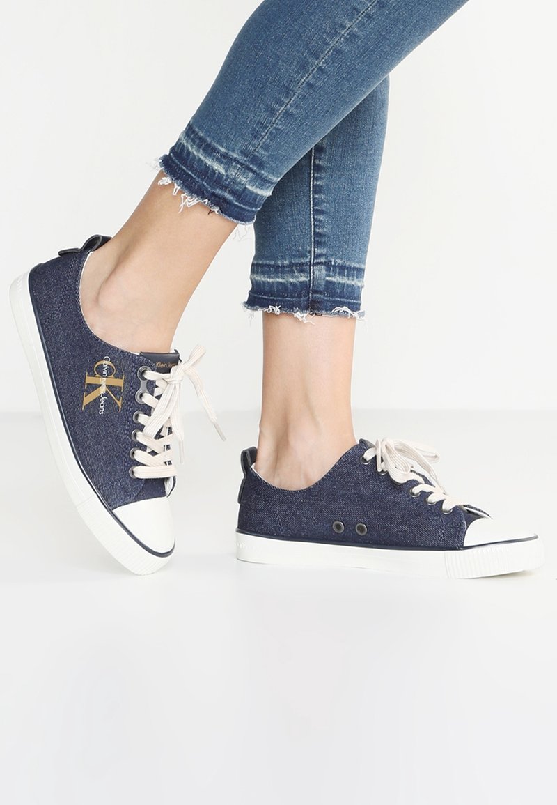 Marinblå denimskor med beige snörning, vit gummikap och logotypdetalj. Har öglor på sidan och en slät sula.