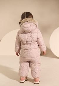 Pufasta ružičasta skijaška odijela s kapuljačom obloženom krznom, ukrašena zlatnim i crnim zvjezdanim uzorcima. Rukavi i nogavice u punoj dužini s elastičnim strukom.