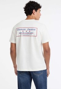 Mężczyzna z kręconymi włosami, ubrany w białą koszulkę z napisem "Guess Jeans Denim Company" oraz niebieskie dżinsy, obrócony, aby pokazać tył koszulki.