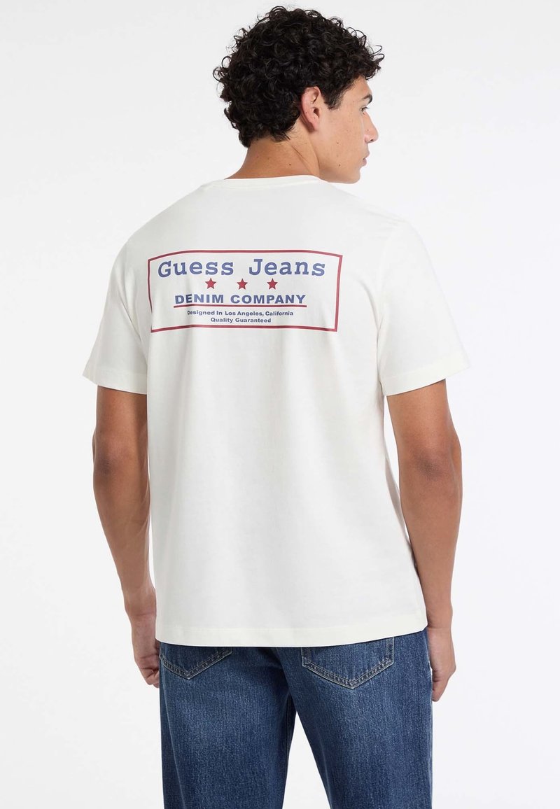 Mężczyzna z kręconymi włosami, ubrany w białą koszulkę z napisem "Guess Jeans Denim Company" oraz niebieskie dżinsy, obrócony, aby pokazać tył koszulki.