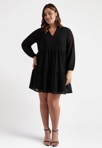 Vestido negro con mangas largas de gasa, escote en V y falda acampanada. Tejido ligero con un diseño fluido, largo hasta la rodilla, combinado con tacones negros.