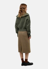 Pull en tricot vert avec un large col, associé à une jupe à carreaux de longueur genou. Des chaussettes brunes et des chaussures noires complètent la tenue.
