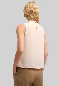 Ärmlös ljusrosa blus med en liten nyckelhålsdetalj på ryggen, slät textur och avslappnad passform, matchad med beiga byxor.