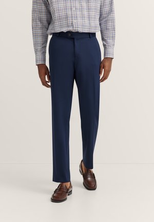 Mann trägt marineblaue Anzughose, braune Loafers und ein langärmliges kariertes Hemd und läuft vor hellem Hintergrund.