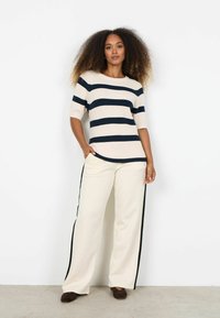 Pruhovaný pletený sveter v krémovej a námorníckej modrej farbe, kombinovaný s krémovými širokými nohavicami so čiernymi bočnými pruhmi. Model stojí s rukou vrecku.