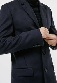 Chaqueta azul marino de mezcla de lana con solapas con muescas, botones abotonados en fila simple y bolsillos laterales. Se aprecia la tela texturizada y el ajuste entallado.