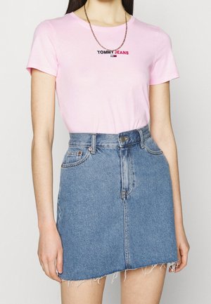 Kobieta ubrana w różowy t-shirt Tommy Jeans i niebieską spódnicę dżinsową z wysokim stanem i postrzępionym obszyciem, uzupełnioną złotym naszyjnikiem z łańcuszkiem.