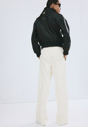 Personne aux cheveux tressés portant une veste en cuir noire, un pantalon blanc à jambes larges et des lunettes de soleil noires, debout les mains dans les poches, tournée de dos.