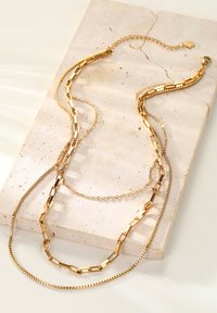 Złote naszyjniki warstwowe o różnych wzorach łańcuchów, w tym pudełkowym, płaskim i delikatnym, leżące na teksturowanej neutralnej powierzchni.