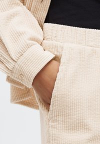 Helle beige Cordjacke mit gerippter Textur, ausgestattet mit einem knöpfbaren Bündchen und einer Hand, die in der Tasche einer passenden Hose steckt.