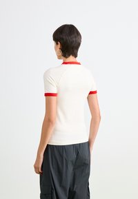 T-shirt blanc à manches courtes avec un col et des poignets rouges, arborant un design ajusté et une texture lisse, associé à un pantalon foncé ample.