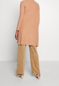 Pfirsichfarbener gerippter langer Cardigan mit geradem Schnitt, kombiniert mit sandfarbenen Corduroy-Hosenträgern und spitzen beigen Absatzschuhen.