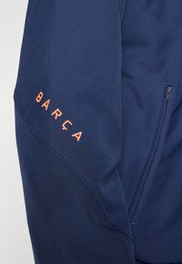 Marineblaue Sportjacke mit orangefarbenem "BARÇA"-Schriftzug. Verfügt über eine glatte Textur, eine Reißverschlusstasche und eine schräg verlaufende Nahtdetail am Ärmel.