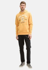 Gelbes Hoodie mit weißem Logo-Print, Kängurutasche und Kapuze mit Zugband. Kombiniert mit schwarzen Hosen und hellgrauen Sneakers.
