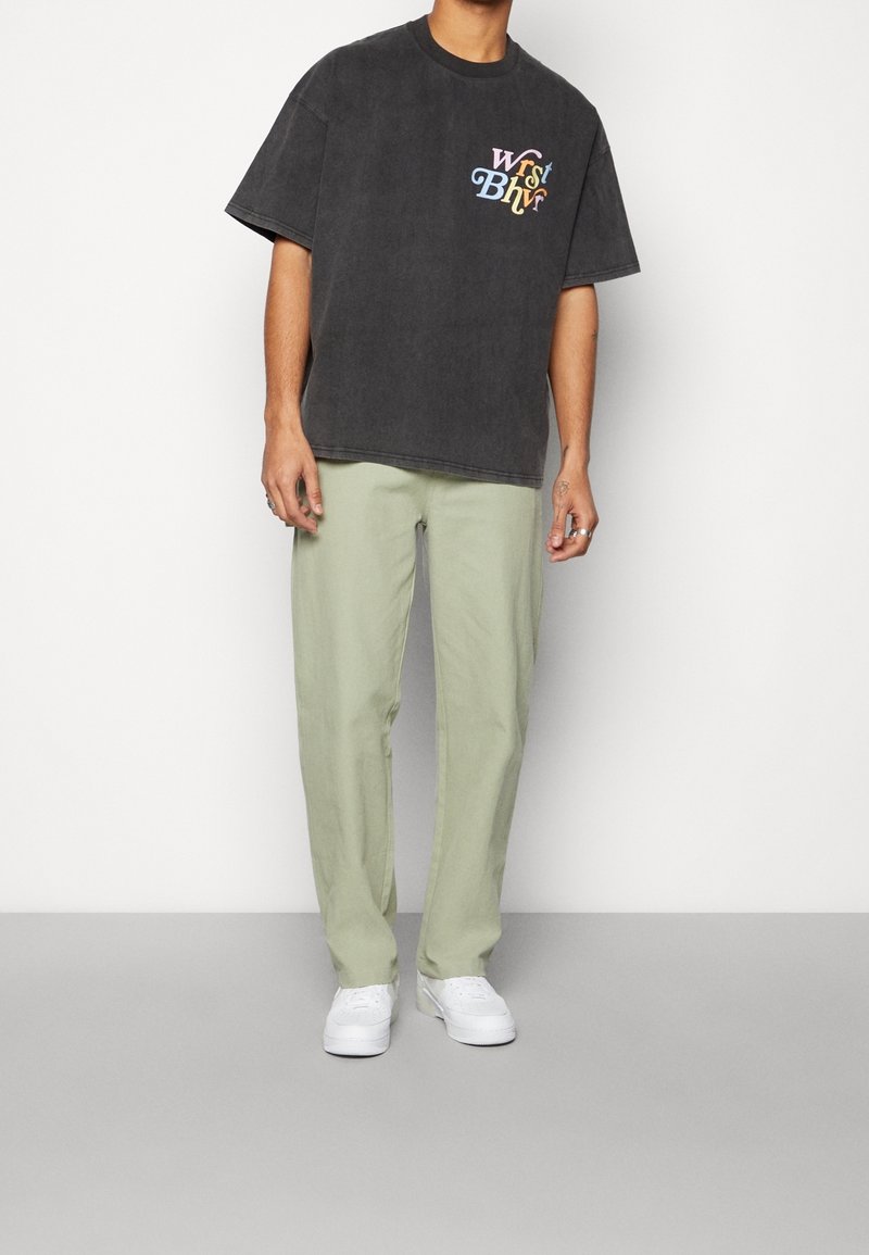 T-shirt gris surdimensionné à manches courtes avec logo multicolore sur la poitrine, assorti à un pantalon droit vert clair et des baskets blanches.