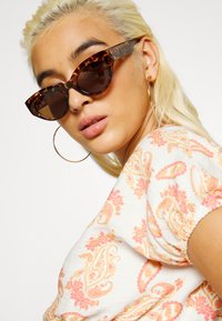 Óculos de sol estilo gato com armação de tartaruga, lentes castanhas e formato oversized. A modelo usa uma blusa estampada com cores pastel e mangas bufantes.