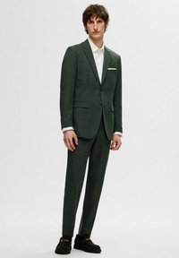 Tumši zaļš pieguļošs uzvalks ar strukturētu audumu, vienrindas žakete ar atvērtām atloks, un saskanīgas bikses. Baltā krekla apakšā, melni apavi.