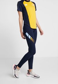 Strój sportowy Adidas z żółtą i granatową górą, granatowymi legginsami z niebieskimi i złotymi akcentami oraz lekkimi wielokolorowymi sneakersami.