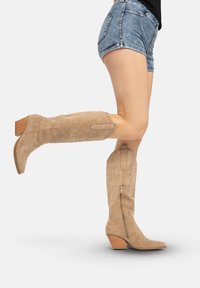 Beige suède kniehoge laarzen met een spitse neus, reliëfpatroon en een zijrits. De hak is laag en lijkt op gestapeld hout.