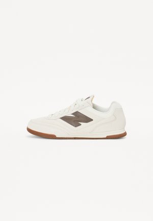 Biały niski sneaker z brązową podeszwą i ciemnobrązowym logo z boku, zawiązywany na sznurówki, z gładkim skórzanym wykończeniem.